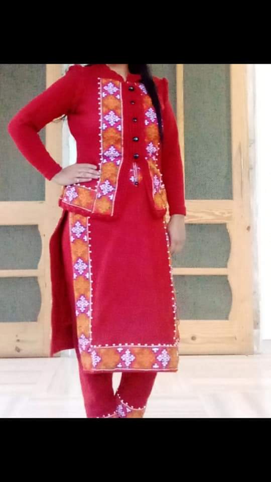 A woman stands indoors wearing a red dress with orange and white KULLU Patti (LACE) border from aruna kullu handloom, paired with matching pants and Pula Chappal. Her hand is on her hip; her face is not visible.