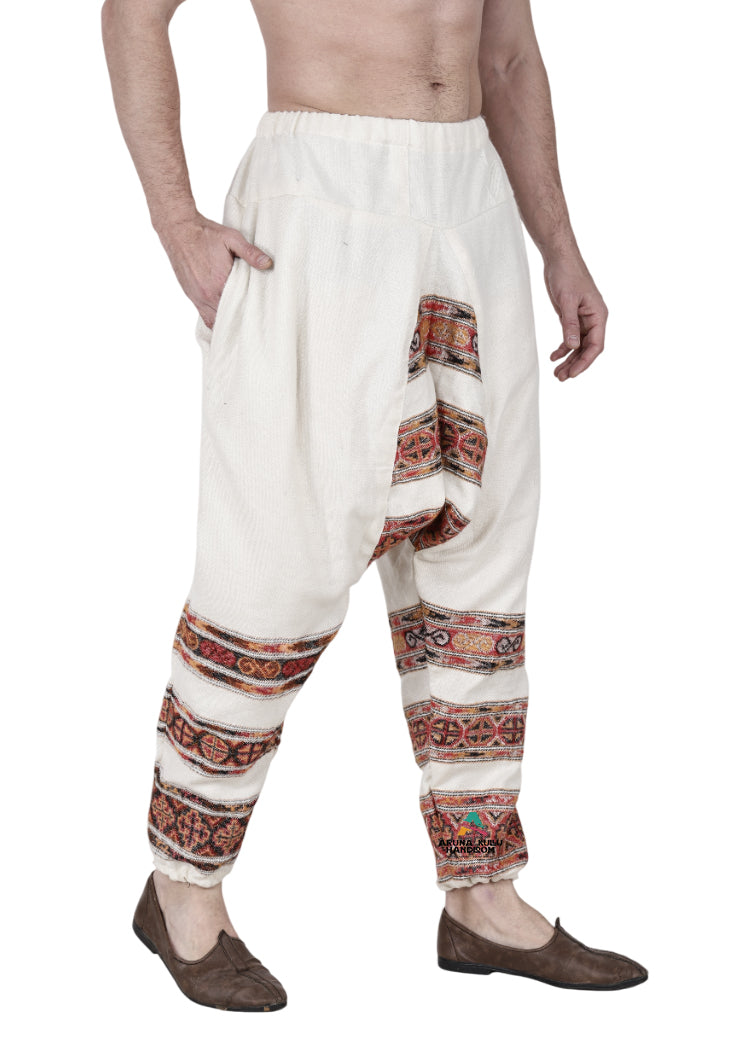 A person wears aruna kullu handloom yoga pajama for men loose white 3 patti with colorful leg embroidery and brown traditional slip-on shoes. The upper body is bare against a white background.