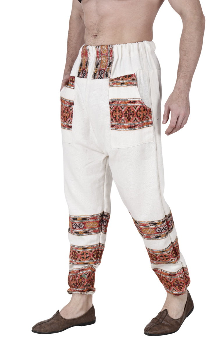 A person wears white "Pajama for men 3 patti" by aruna kullu handloom, featuring hand-woven embroidery, colorful patterns, elastic waistband, large front pockets, paired with brown slip-on shoes; model’s torso not fully visible.