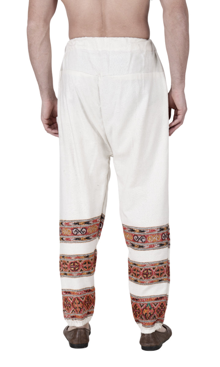 A person is seen from the back wearing aruna kullu handloom’s Pajama for men white 3 patti, featuring colorful handwoven embroidery around the lower legs and traditional shoes, with a bare upper body.