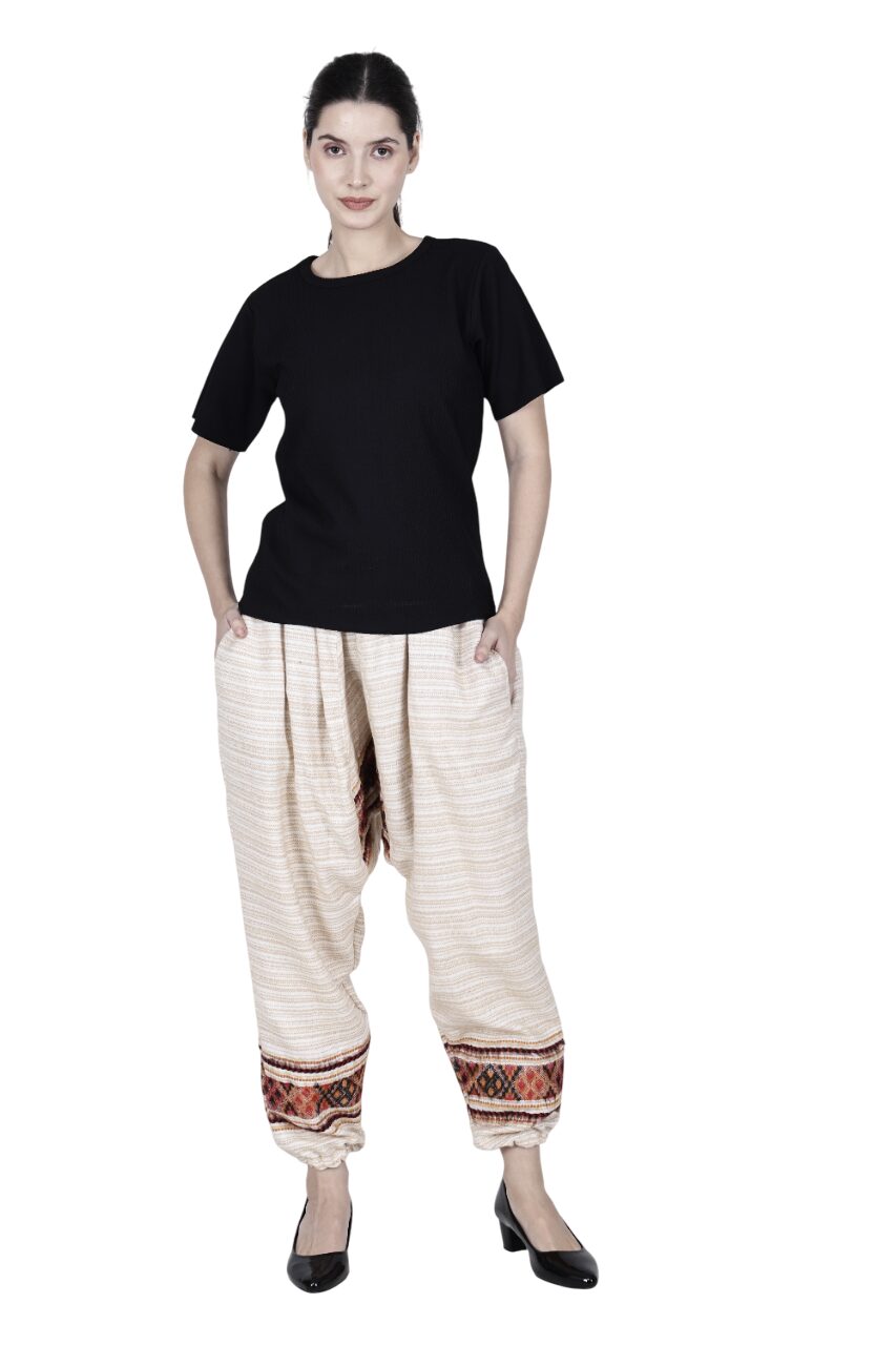 A woman faces forward, wearing Aruna Kullu Handloom's loose yoga pajama "white singal patti," cream-colored with orange and black patterned cuffs. She has her hands in her pockets and wears a black short-sleeve top and black shoes.