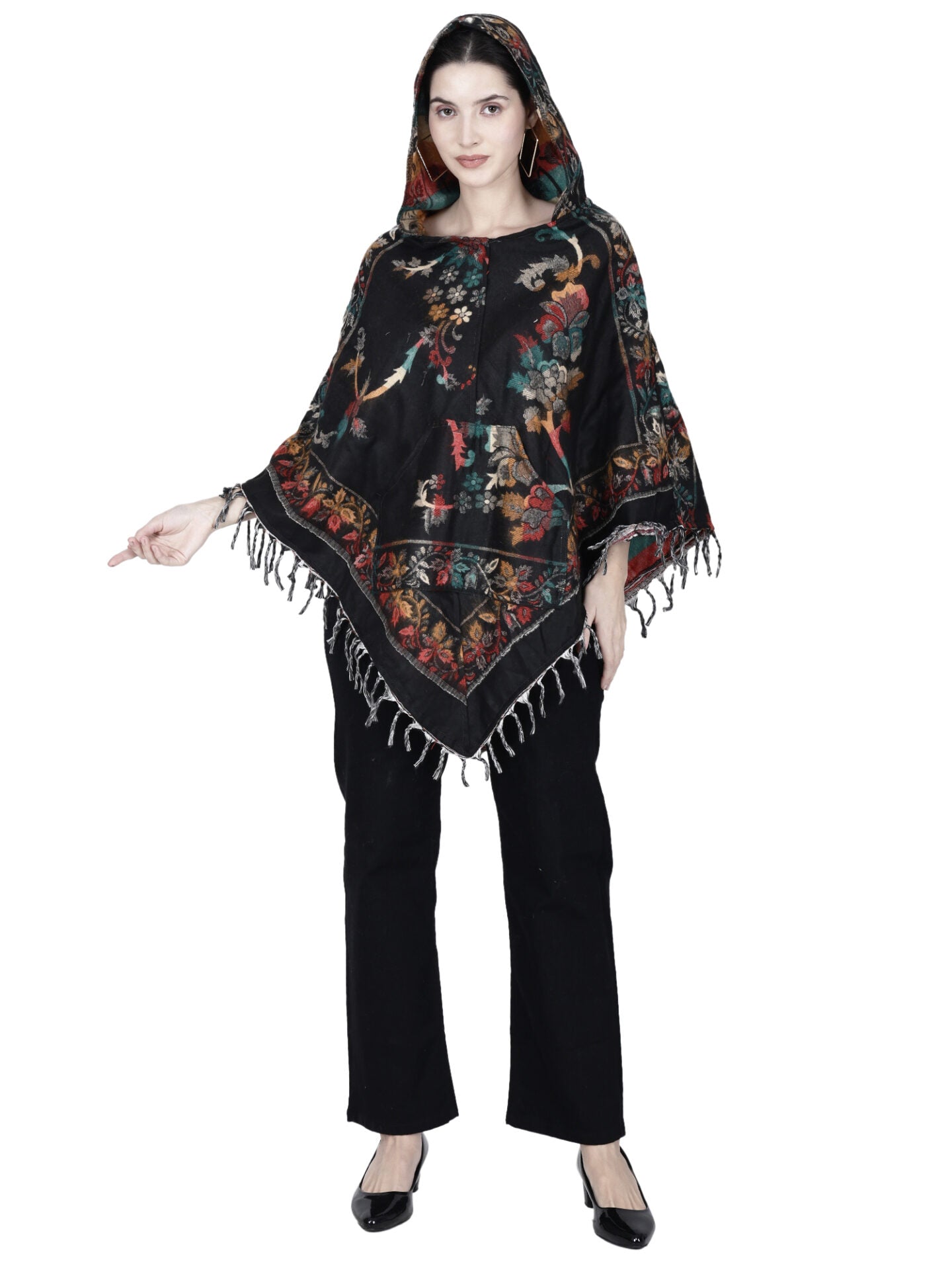 A woman models the aruna kullu handloom Kashmiri woolen poncho in black, featuring a hood, colorful floral patterns, and fringe details, paired with black pants and flats. She stands with one hand slightly raised.