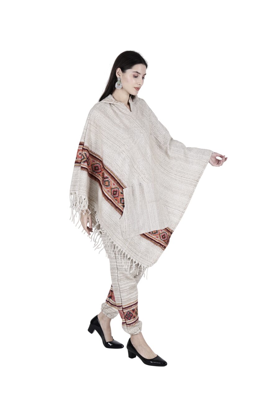 A woman wears the Aruna Kullu Handloom "combo pajama and poncho singal patti white," featuring a handwoven beige poncho with red and black geometric designs, matching pants, black heels, and bold earrings against a white background.