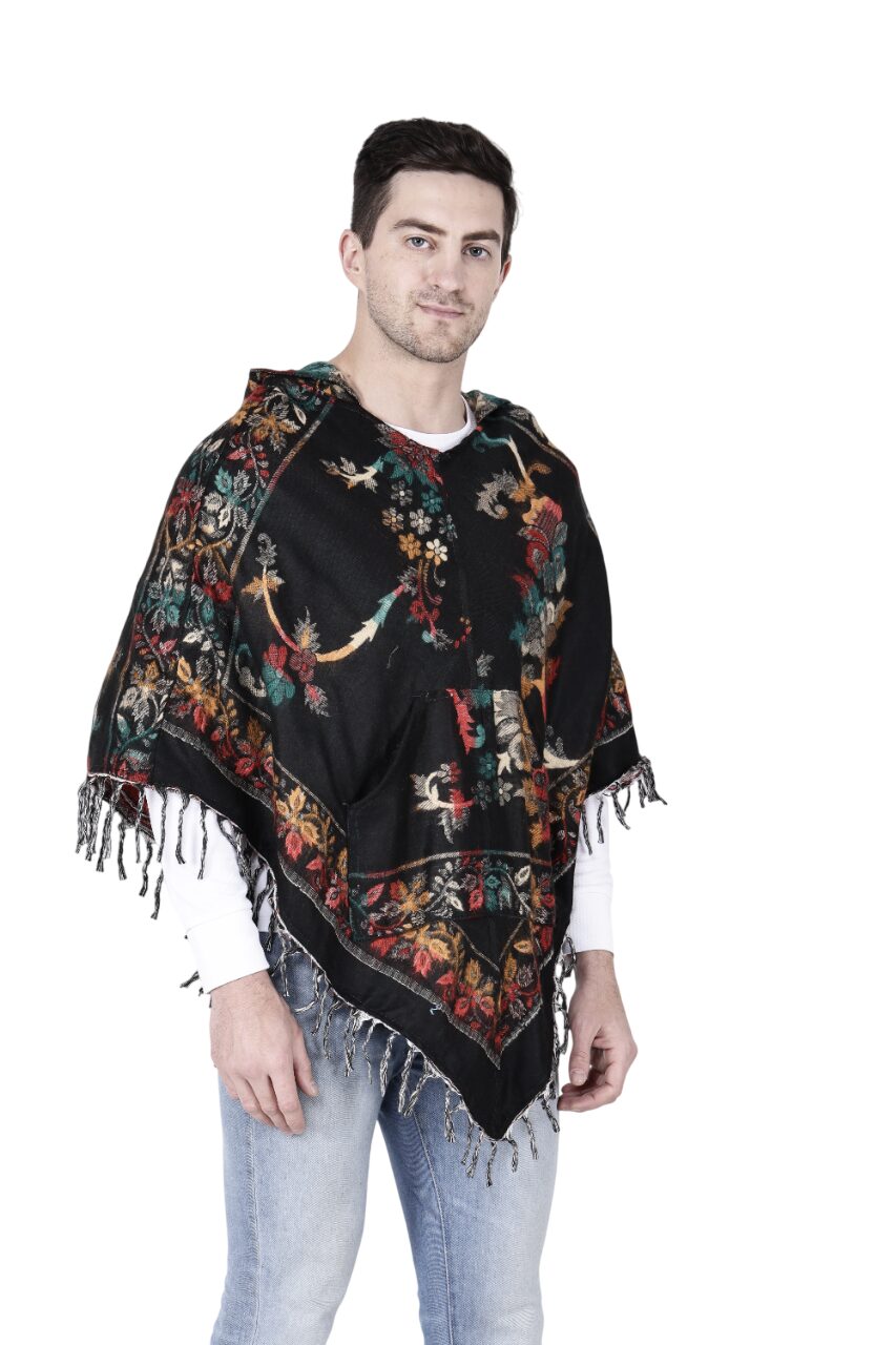 A man stands against a white background wearing the aruna kullu handloom Kashmiri poncho black, featuring colorful floral embroidery and fringe details. He pairs the handwoven piece with a white long-sleeve shirt and light blue jeans.