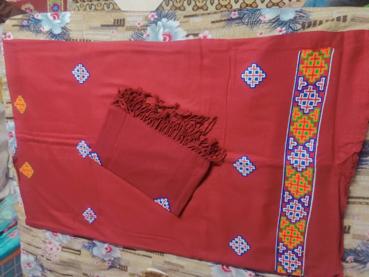 Aruna Kullu Handloom's "Kullu Patti Woolen Suit 4 Meter with Stole" in maroon features traditional geometric embroidery in blue, yellow, red, and green motifs along the edge; includes a matching fringed stole and is set against a floral bedsheet.