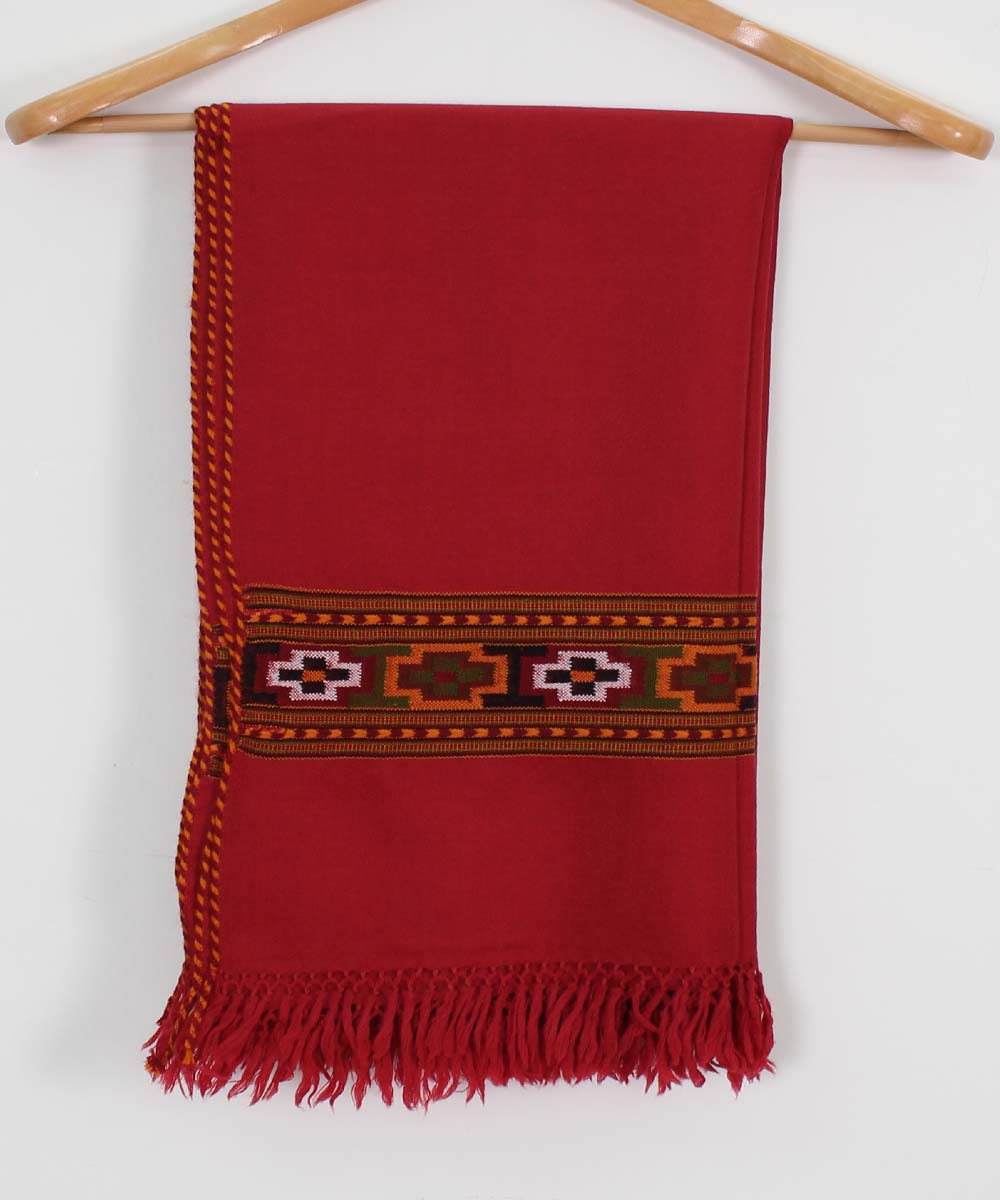 The Kullu Kinnauri Handcrafted Shawl by aruna kullu handloom is a pure wool, handwoven women's shawl in red with black, white, and brown geometric embroidery near one edge and fringed ends, shown hanging on a wooden hanger.