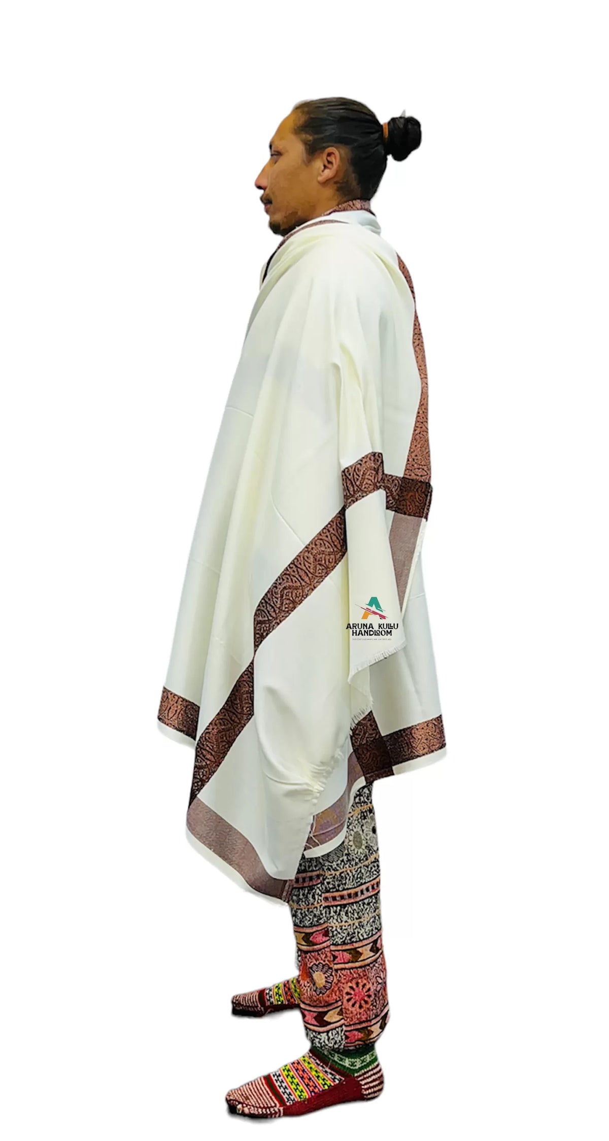 A man stands in profile against a white background, wearing traditional attire including the aruna kullu handloom Genuine Hand Woven pashmina Wool LOI (MEN SHAWL) in white, patterned pants, and colorful beaded moccasins. His hair is tied in a bun.