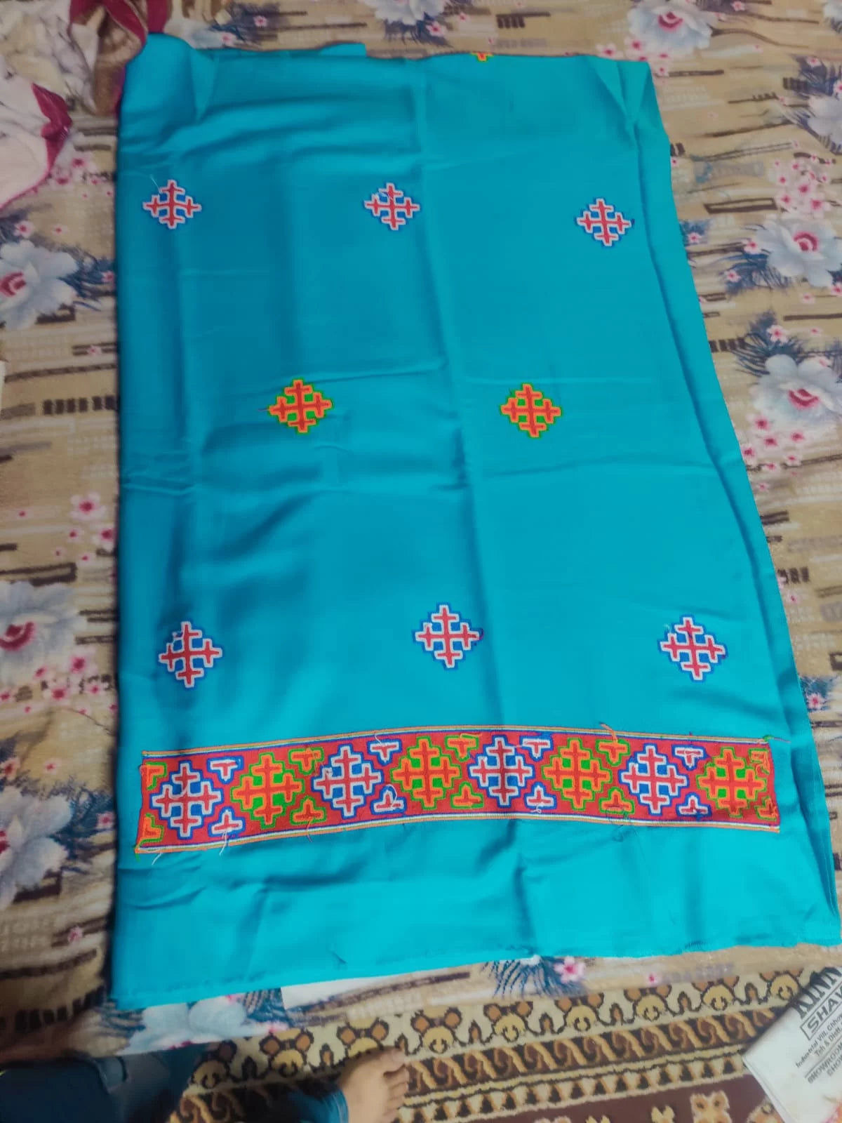 The aruna kullu handloom kullu patti woolen suit (4 meters) with stole in frozen turquoise features colorful geometric embroidery and a wide kullu patti border, displayed on a floral-patterned bedspread.