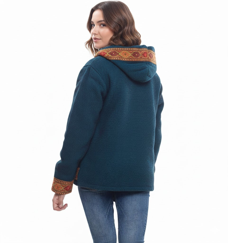 A woman with wavy brown hair wears the aruna kullu handloom Himachal handicrafts Woolen Winter Wear Zipper Hoodie Jacket for Men & Women with blue Kullu Patti work, featuring traditional patterned trim on the hood and cuffs, paired with blue jeans.himachal products online himachali products online bhuttico online shopping himachal handloom online himachal products online shopping
