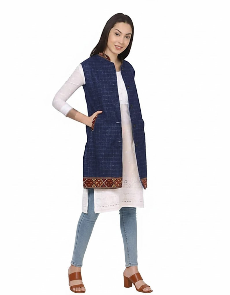 A woman smiles, wearing a long white kurta, aruna kullu handloom's check navy blue long jacket with kullu lace work, blue jeans, and brown heeled sandals, posing against a plain white background.half jacket for women khadi half jacket for ladies ladies half jacket cotton women's half jacket ladies sadri design winter half jacket for women