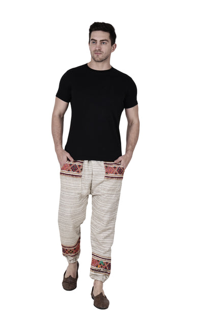 A man in a black t-shirt, wearing "Pajama for men white singal patti" by aruna kullu handloom, featuring colorful handwoven embroidery on the waistband and cuffs, stands with his hands in his pockets against a white background.