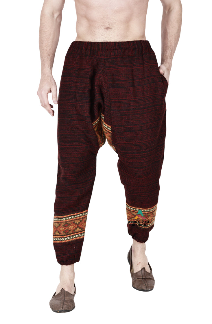 A man stands barefoot, hands in pockets, wearing aruna kullu handloom’s yoga pajama for men in loose maroon style. The pants have colorful ethnic bands near the ankles; his upper body is unclothed.