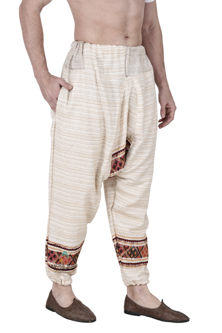 A man models the Aruna Kullu Handloom yoga pajama for men loose single patti white, featuring woven ethnic patterns by the ankles, standing barefoot with one hand in his pocket against a white background.