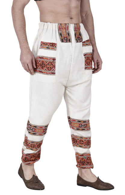 A person wears the "Pajama for men white 3 patti" by aruna kullu handloom, featuring colorful embroidered patterns on the pockets and legs. The upper body is bare, and brown shoes are worn against a white background.