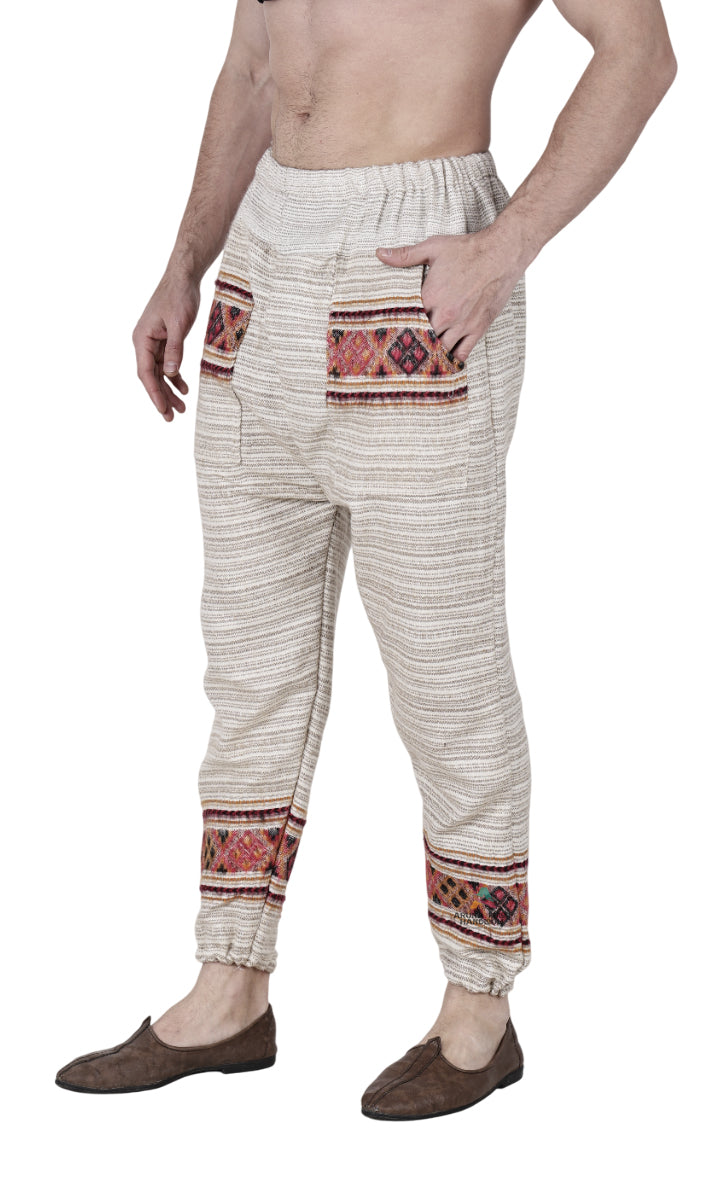 A person wears aruna kullu handloom’s "Pajama for men white singal patti," cream-colored, loose woolen pajamas with elastic waist/ankles and patterned red, orange, black geometric accents and handwoven embroidery, paired with brown slip-ons.
