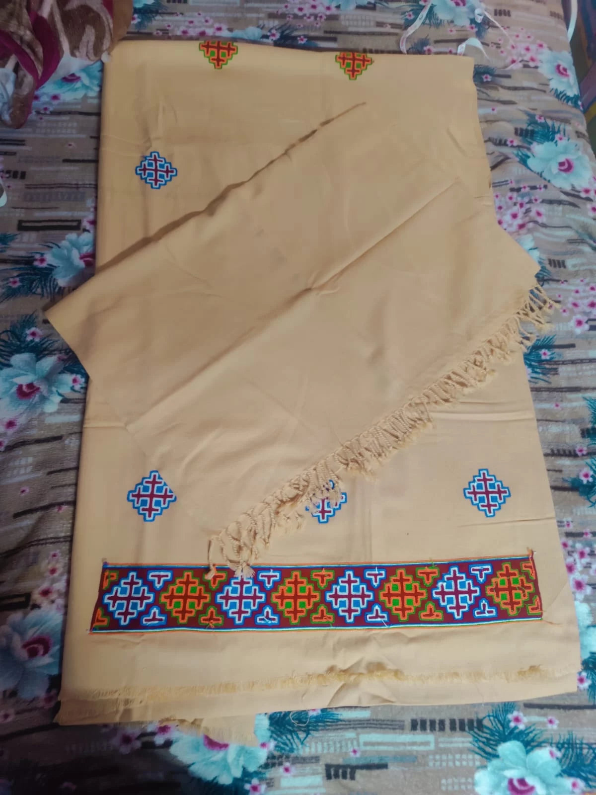 A cream woolen handloom suit by aruna kullu handloom, featuring colorful geometric embroidery and a kullu patti border, is neatly folded with a fringed edge. This 4-meter suit comes with a stole, reflecting classic kullu designs.