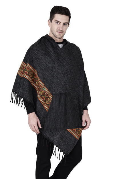 A man stands against a white background wearing a black kullu shawl woolen poncho with hood, featuring a colorful patterned stripe and fringed edges.