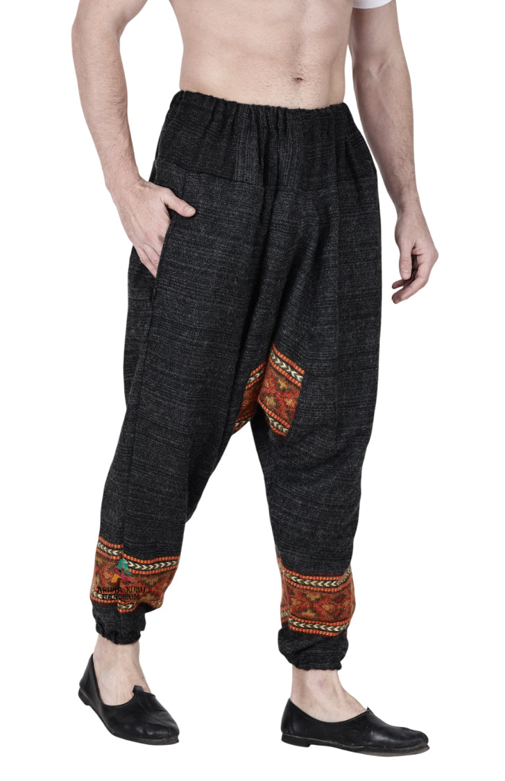 A man stands barefoot against a white background, wearing Aruna Kullu Handloom's yoga pajama for men—loose, black single patti pants with colorful orange and red bands near the ankles and handwoven details.