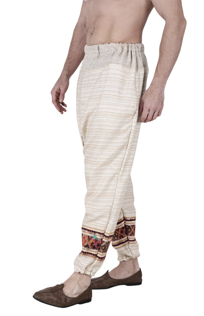 A man wears an Aruna Kullu Handloom yoga pajama for men, loose single patti white, featuring colorful kullu patti embroidery near the ankles and an elastic waistband, paired with brown slip-on shoes. His upper body is bare and partially visible.
