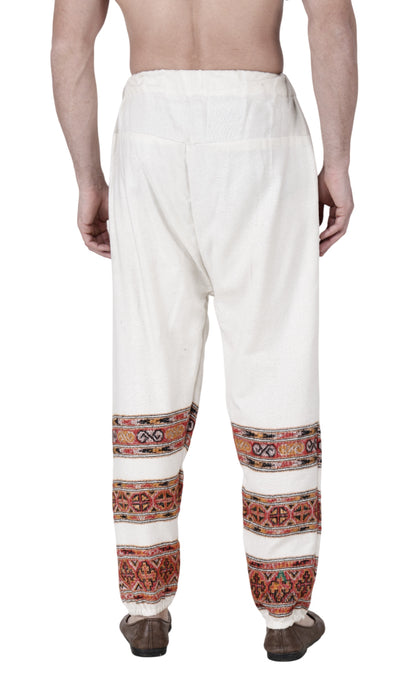 A person is seen from the back wearing aruna kullu handloom’s Pajama for men white 3 patti, featuring colorful handwoven embroidery around the lower legs and traditional shoes, with a bare upper body.