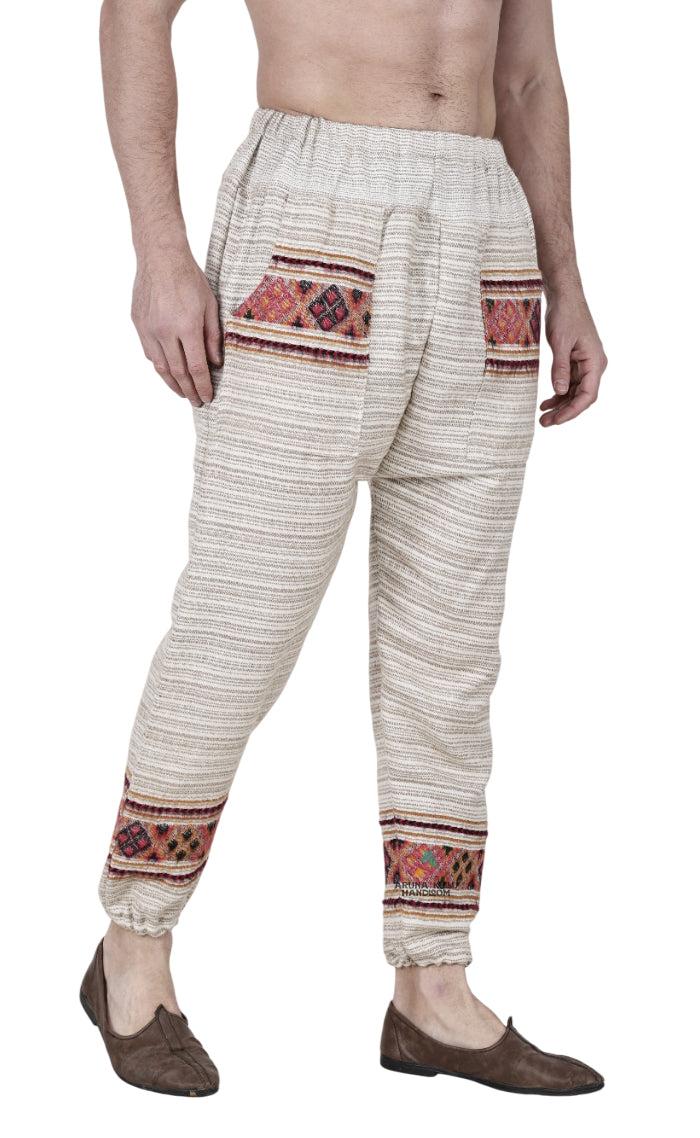 A barefoot man wears aruna kullu handloom's "Pajama for men white singal patti," a traditional white, loose-fitting pajama with colorful handwoven embroidery on the pockets and ankles, against a plain white background.