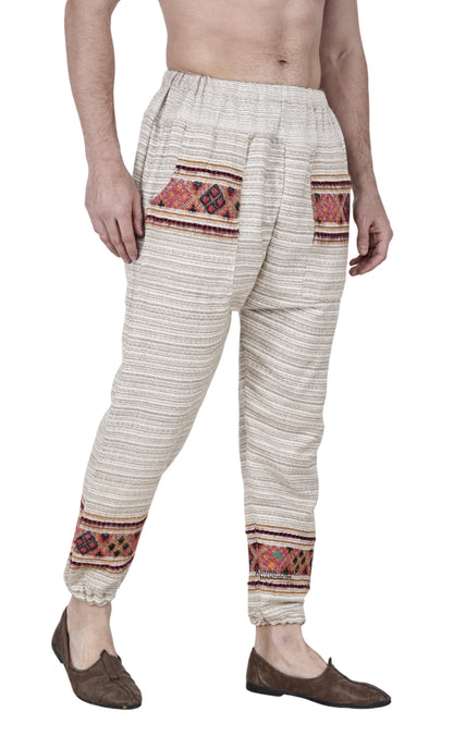 A barefoot man wears aruna kullu handloom's "Pajama for men white singal patti," a traditional white, loose-fitting pajama with colorful handwoven embroidery on the pockets and ankles, against a plain white background.