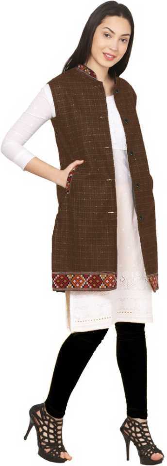 A woman smiles and poses against a white background in an aruna kullu handloom check brown long jacket with kullu lace work, worn over a white dress with black leggings and black high-heeled sandals.
