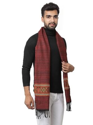 A man in a black turtleneck and white pants models the aruna kullu handloom Kullu muffler—woollen, hand-woven, pure embroidered, and maroon with decorative borders and tasseled ends—smiling as he holds one end of the scarf.