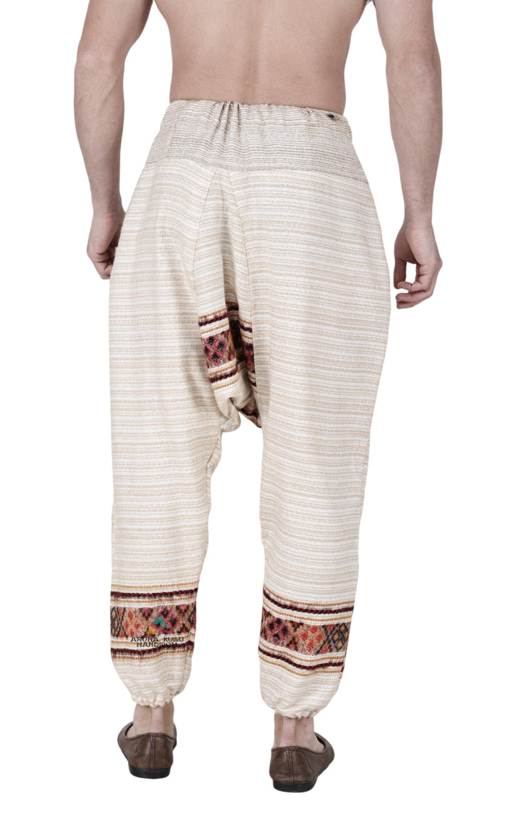 A person stands barefoot, seen from behind, wearing Aruna Kullu Handloom's yoga pajama for men—loose, single patti, white—with colorful woven patterns at the pockets and ankles; their upper body is unclothed.