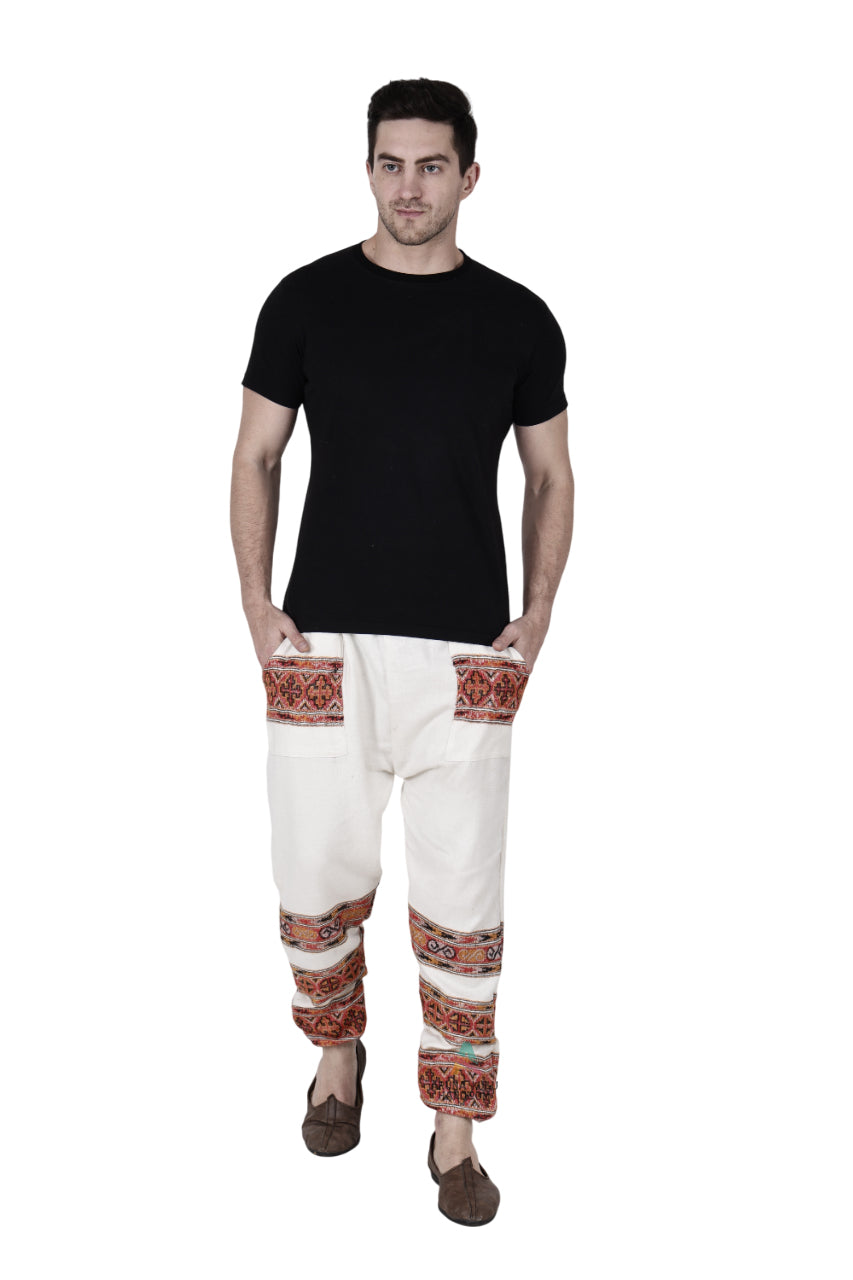A man models the "Pajama for men white 3 patti" by aruna kullu handloom, featuring colorful hand-woven embroidery on the pockets and legs, paired with a black t-shirt, standing with his hands in his pockets against a white background.