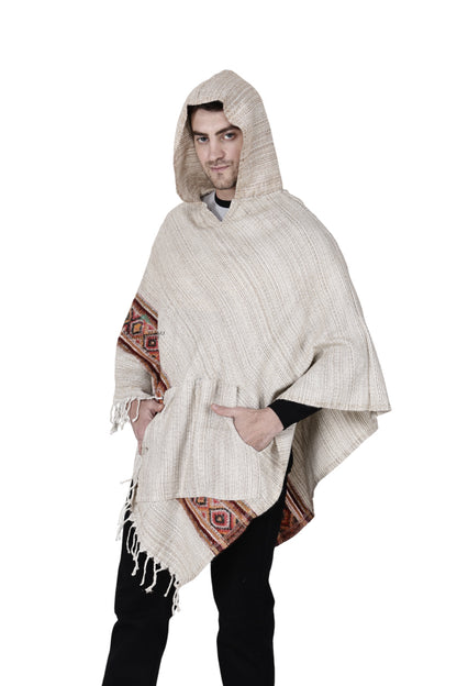 Someone wears a kullu shawl woolen poncho with hood in white, featuring decorative patterns and hand-woven embroidery, standing with hands in pockets against a white background.