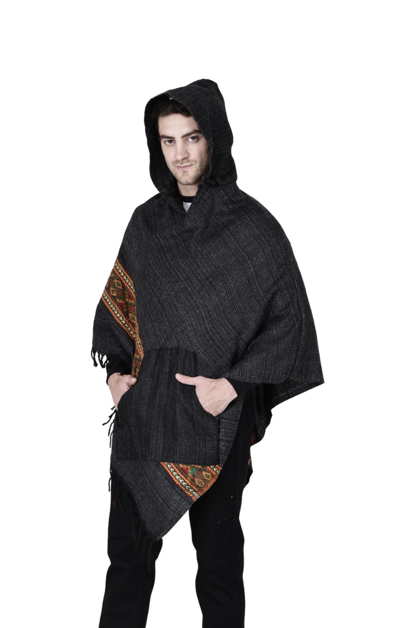 A person models the aruna kullu handloom kullu shawl woolen poncho with hood in black, featuring colorful handwoven embroidery, standing with one hand in the front pocket against a plain white background.
