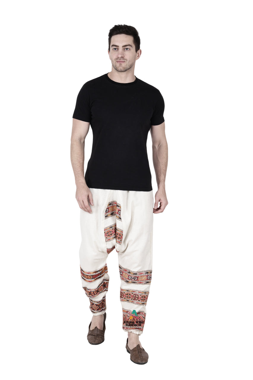 A man stands against a white background wearing a plain black t-shirt, aruna kullu handloom yoga pajama for men loose white 3 patti with colorful ethnic patterns, and brown slip-on shoes.