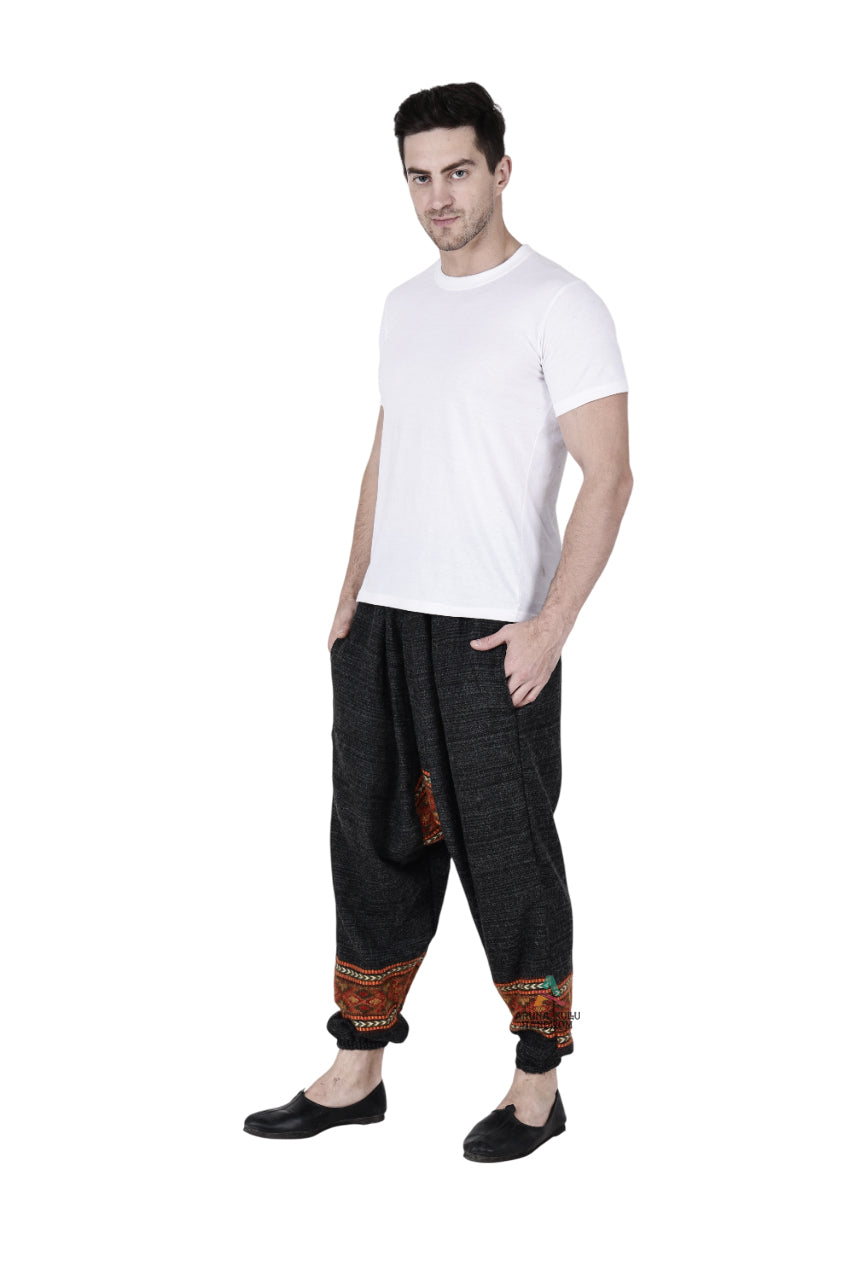 A man poses against a white background, wearing an Aruna Kullu Handloom yoga pajama for men—loose fit with black single patti and handwoven embroidery near the cuffs—paired with a plain white t-shirt and black slip-on shoes.