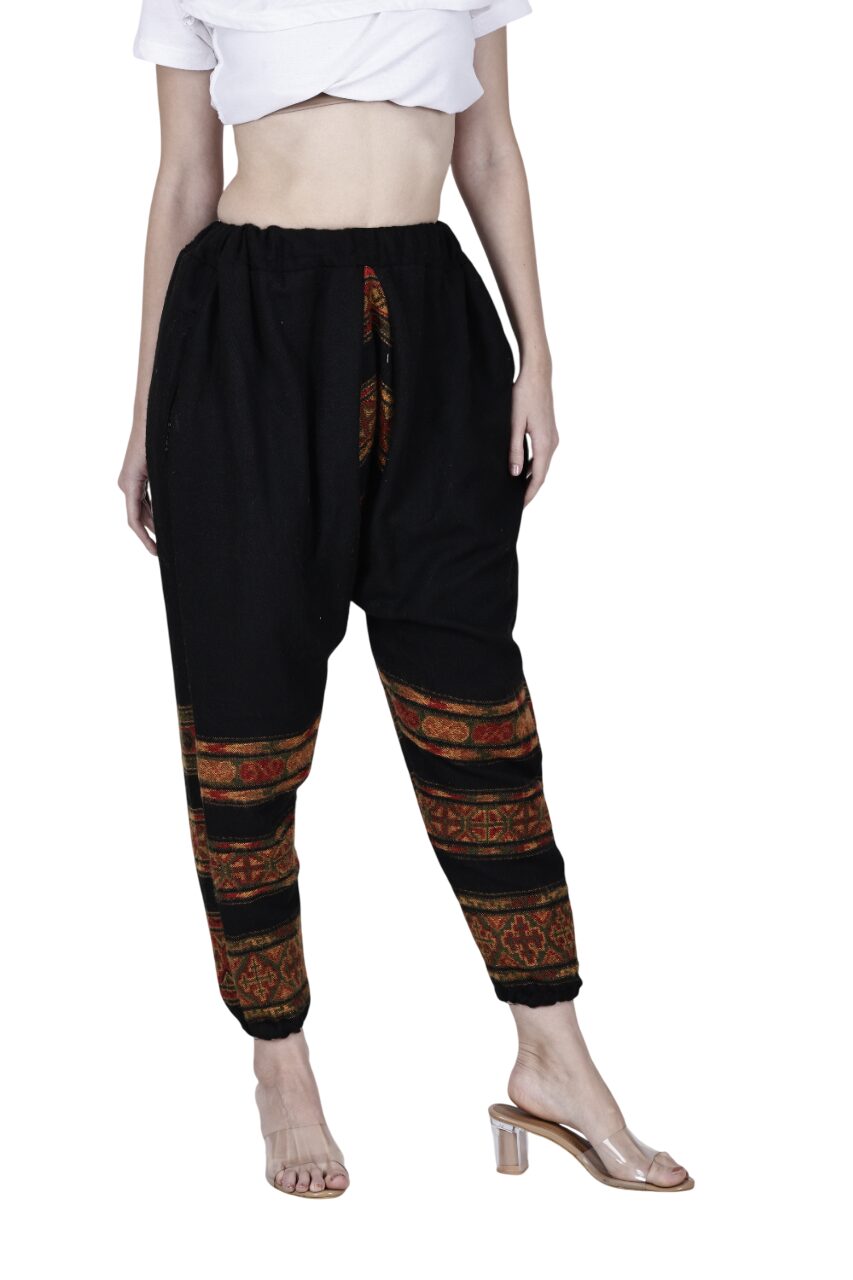 A person wearing aruna kullu handloom yoga loose pajama black with colorful lower leg patterns, paired with a white crop top and clear-heeled sandals, stands against a white background.