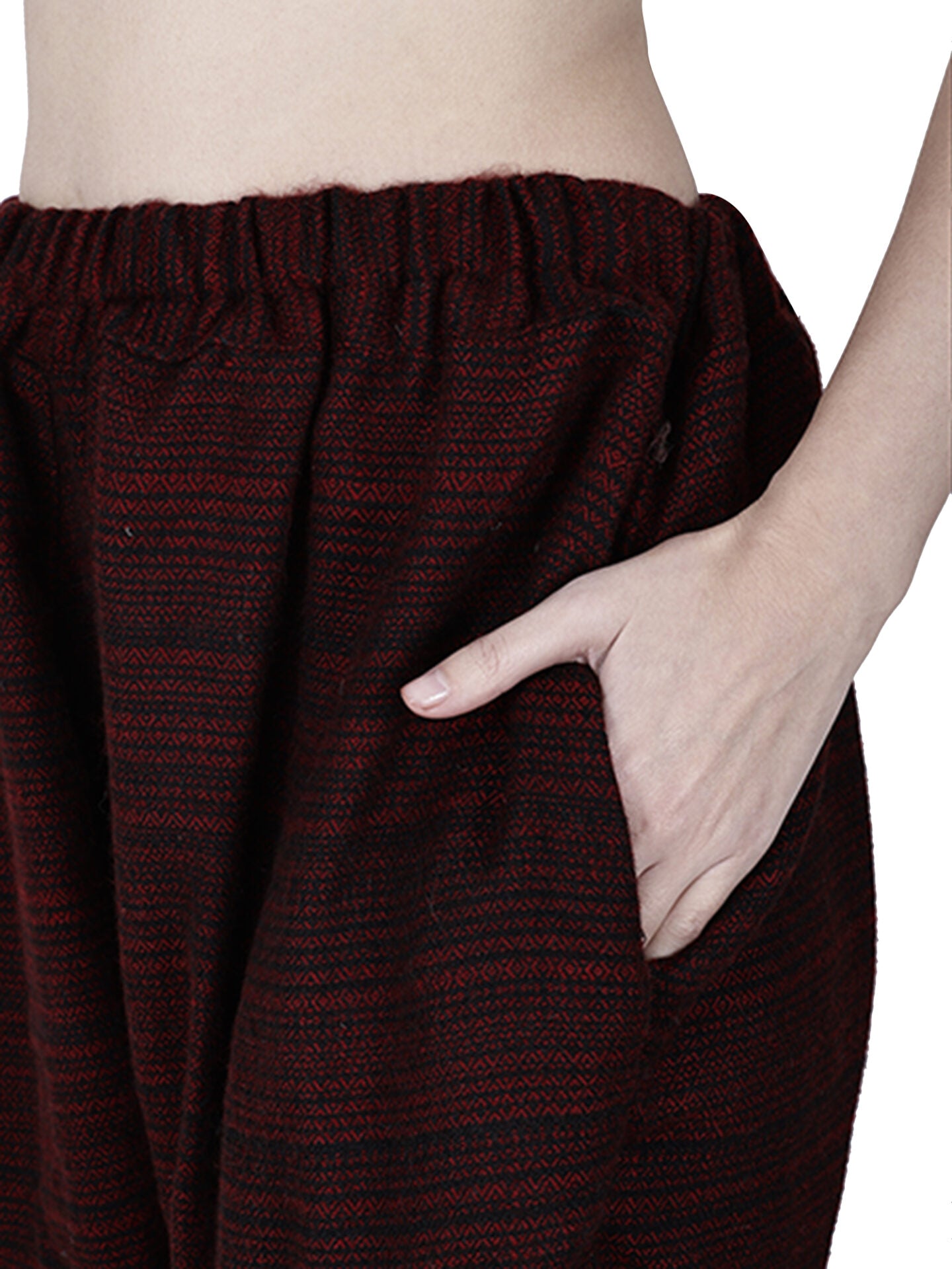 A person wears loose yoga pajama maroon singal patti by aruna kullu handloom—dark red and black patterned woolen pants with elastic waistband, shown waist down against a white background, one hand in pocket. Ideal winter lower for men.