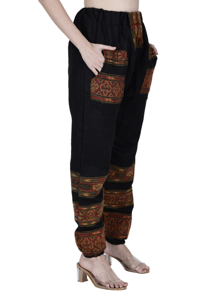 A person wearing aruna kullu handloom’s "winter pajamas black 3 patti" with red and gold handwoven embroidery on the pockets and lower legs, paired with clear high-heeled sandals. The upper body is not visible.