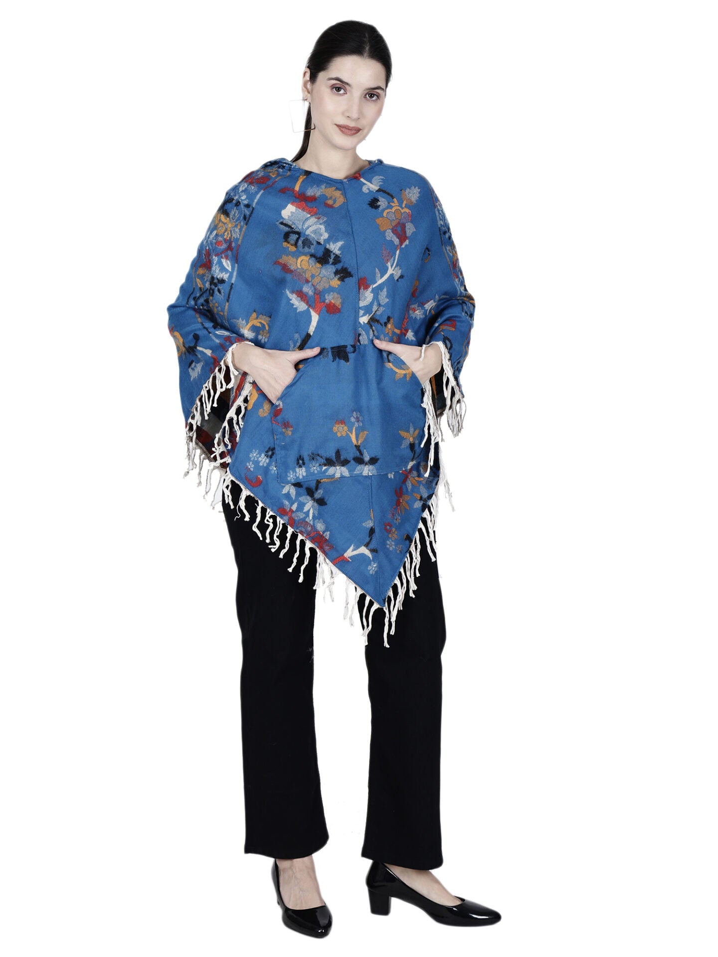 A woman with dark hair tied back stands against a white background, hands in pockets, wearing the aruna kullu handloom Kashmiri poncho woolen poncho with hood for ladies in blue, styled with black pants and black heels.