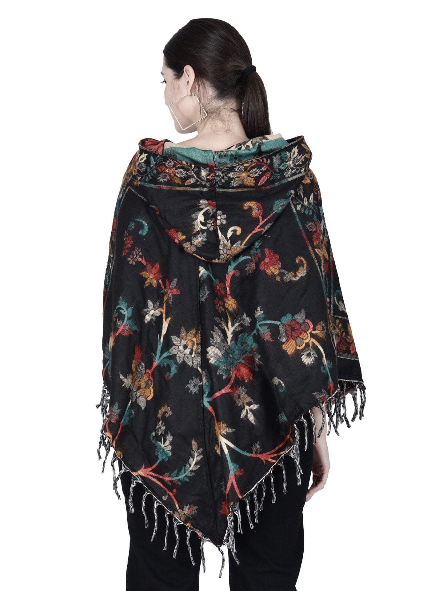 A woman with dark hair in a ponytail is seen from behind wearing the aruna kullu handloom Kashmiri poncho woolen poncho with hood for ladies in black, featuring colorful floral patterns and fringe.