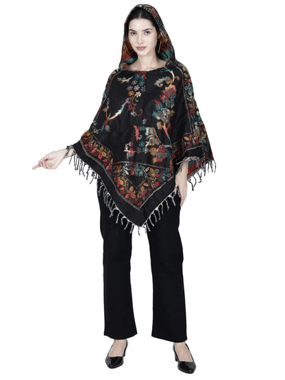 A woman models the aruna kullu handloom Kashmiri woolen poncho in black, featuring a hood, colorful floral patterns, and fringe details, paired with black pants and flats. She stands with one hand slightly raised.