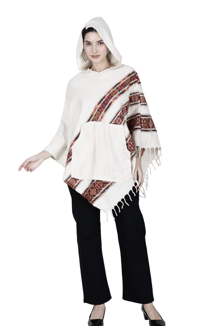 A woman models the Aruna Kullu Handloom woolen poncho with hood, "Kullu Shawl White 3 Patti," featuring red and brown geometric patterns and fringed edges, paired with black pants and shoes, against a white background.