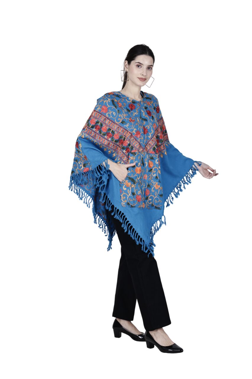 A woman wears the aruna kullu handloom kashmiri pheran—a Kashmiri embroidered woolen poncho with hood for ladies frozen—featuring colorful floral patterns and fringe, paired with black pants and black heels against a white background.