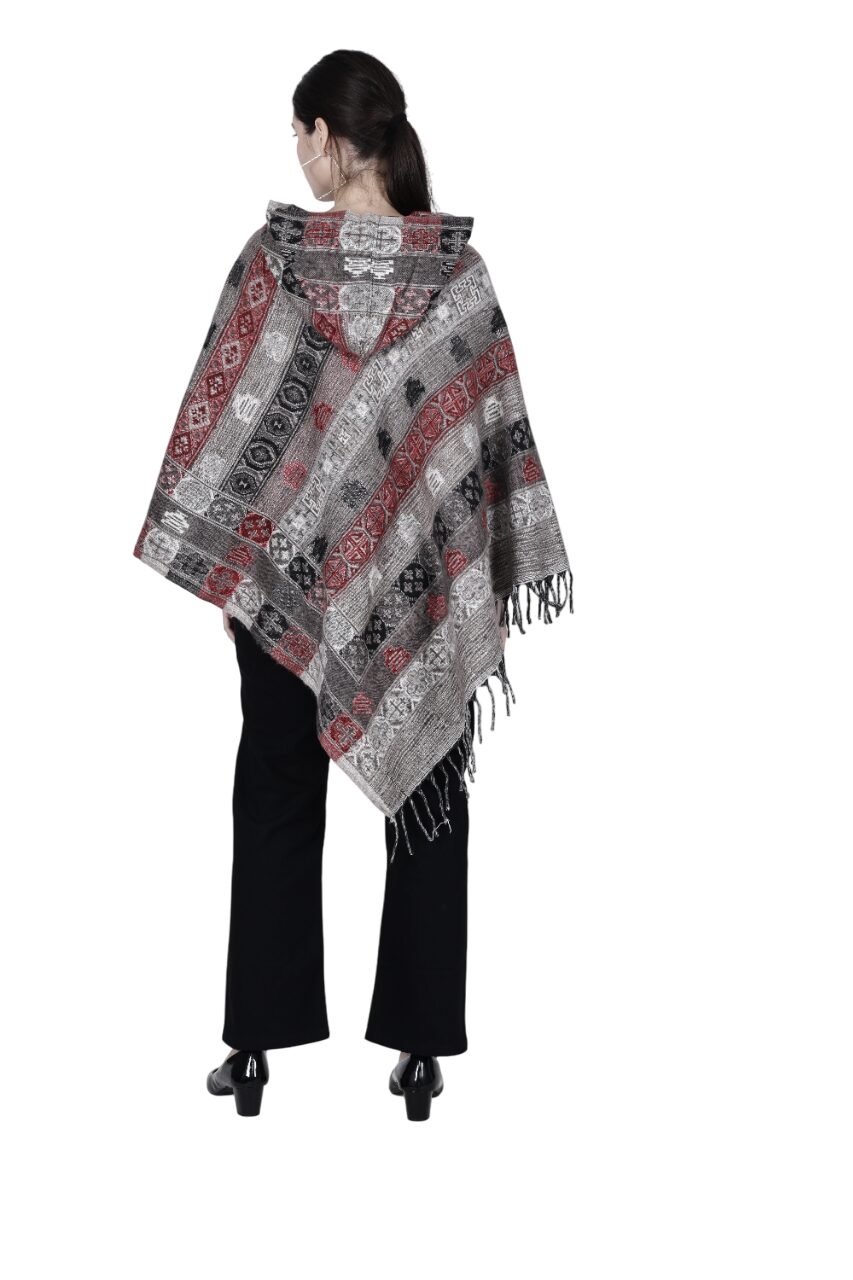 A woman with long brown hair, in black pants and heeled shoes, models the aruna kullu handloom Aruna Kullu handloom shawl poncho for women and girls. The poncho has red, black, and gray geometric patterns with fringe. White background.