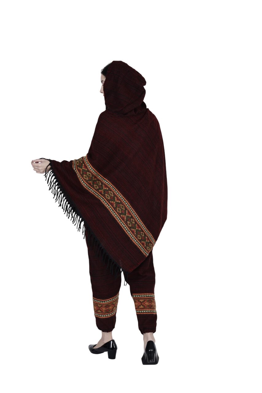 A person in aruna kullu handloom’s combo pajama and poncho singal patti maroon, featuring colorful geometric patterns and hand-woven embroidery, stands turned away against a white background, holding out the fringed edge.