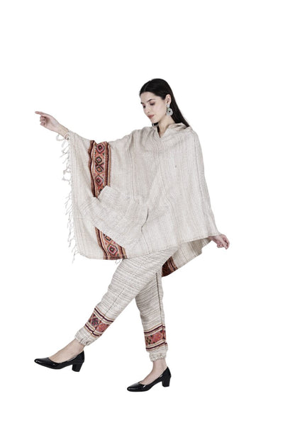 A woman models the "combo pajama and poncho singal patti white" by Aruna Kullu Handloom—light beige, textured two-piece with red and brown borders and a Himalayan poncho—styled with black heels and statement earrings against a white background.