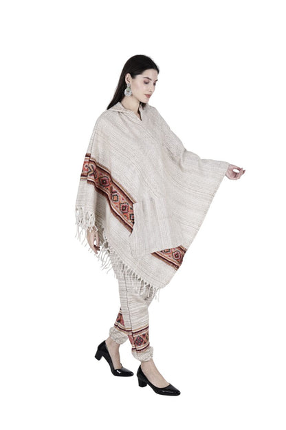 A woman wears the Aruna Kullu Handloom "combo pajama and poncho singal patti white," featuring a handwoven beige poncho with red and black geometric designs, matching pants, black heels, and bold earrings against a white background.