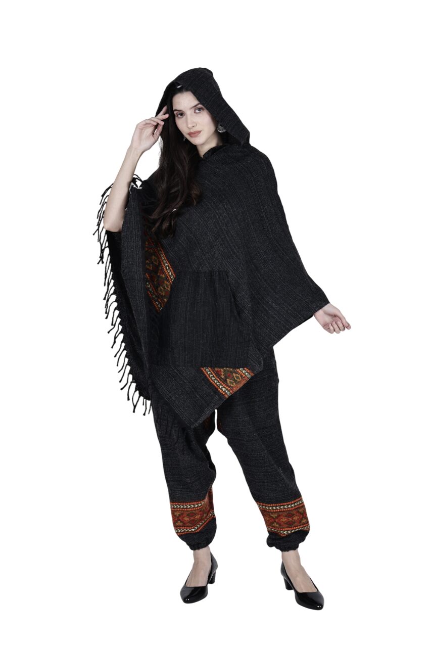 A woman wears the "combo pajama and poncho black singal patti black" by aruna kullu handloom—featuring a dark poncho with fringe and pocket, matching loose pants with patterned cuffs, and black heels—against a white background.