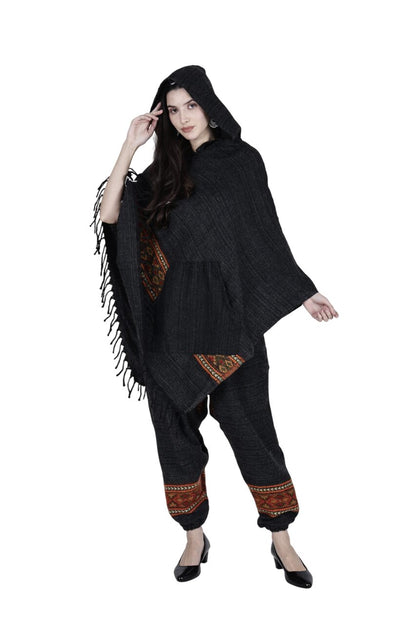 A woman wears the "combo pajama and poncho black singal patti black" by aruna kullu handloom—featuring a dark poncho with fringe and pocket, matching loose pants with patterned cuffs, and black heels—against a white background.