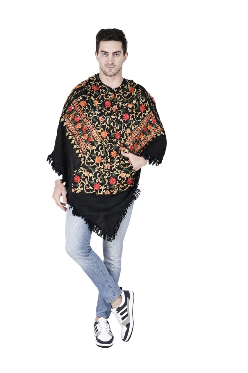 A man wearing an aruna kullu handloom poncho kashmiri embroidery black, light blue jeans, and white sneakers stands with one leg crossed, smiling at the camera against a white background.