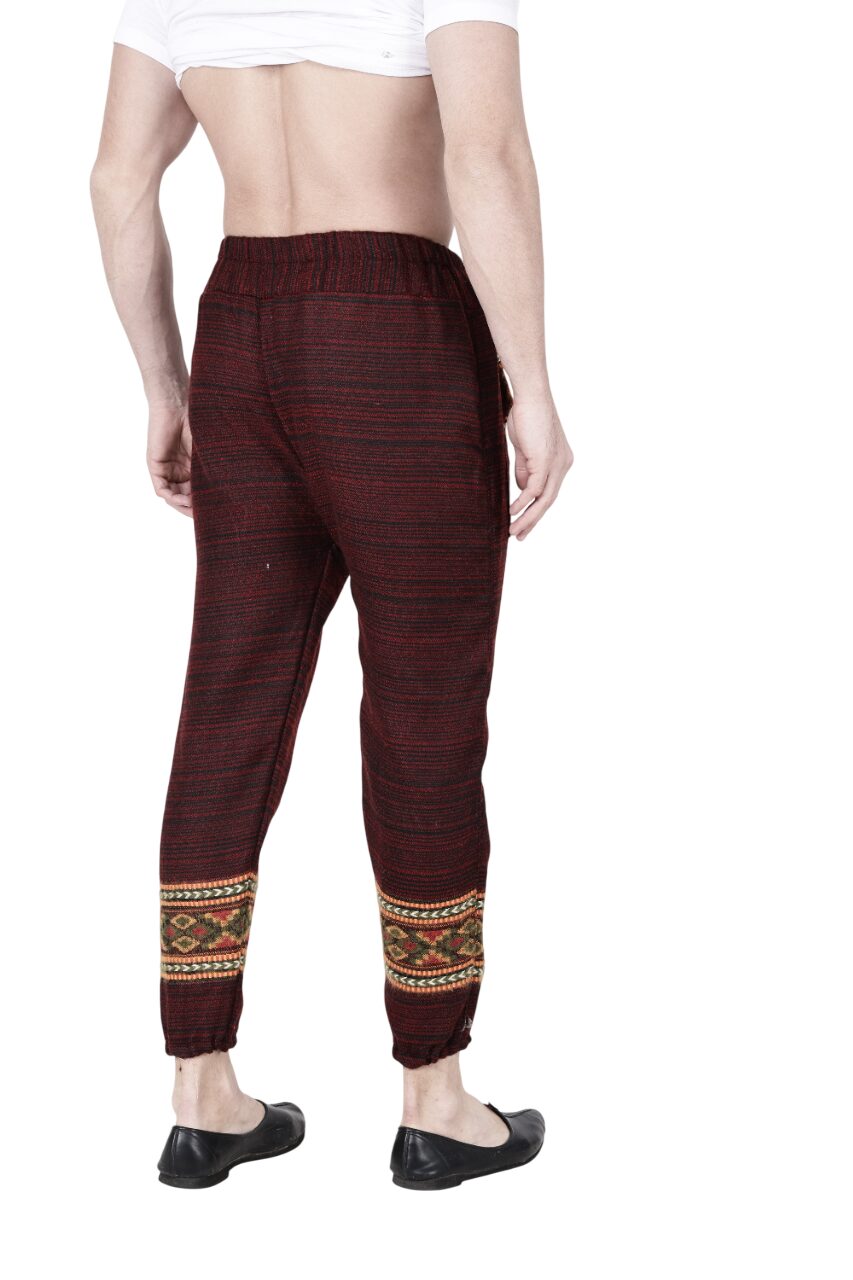 A person faces away from the camera, showcasing aruna kullu handloom's "Pajama for men maroon" with patterned wool and ankle bands, styled with a white cropped shirt and black slip-on shoes against a plain white backdrop.